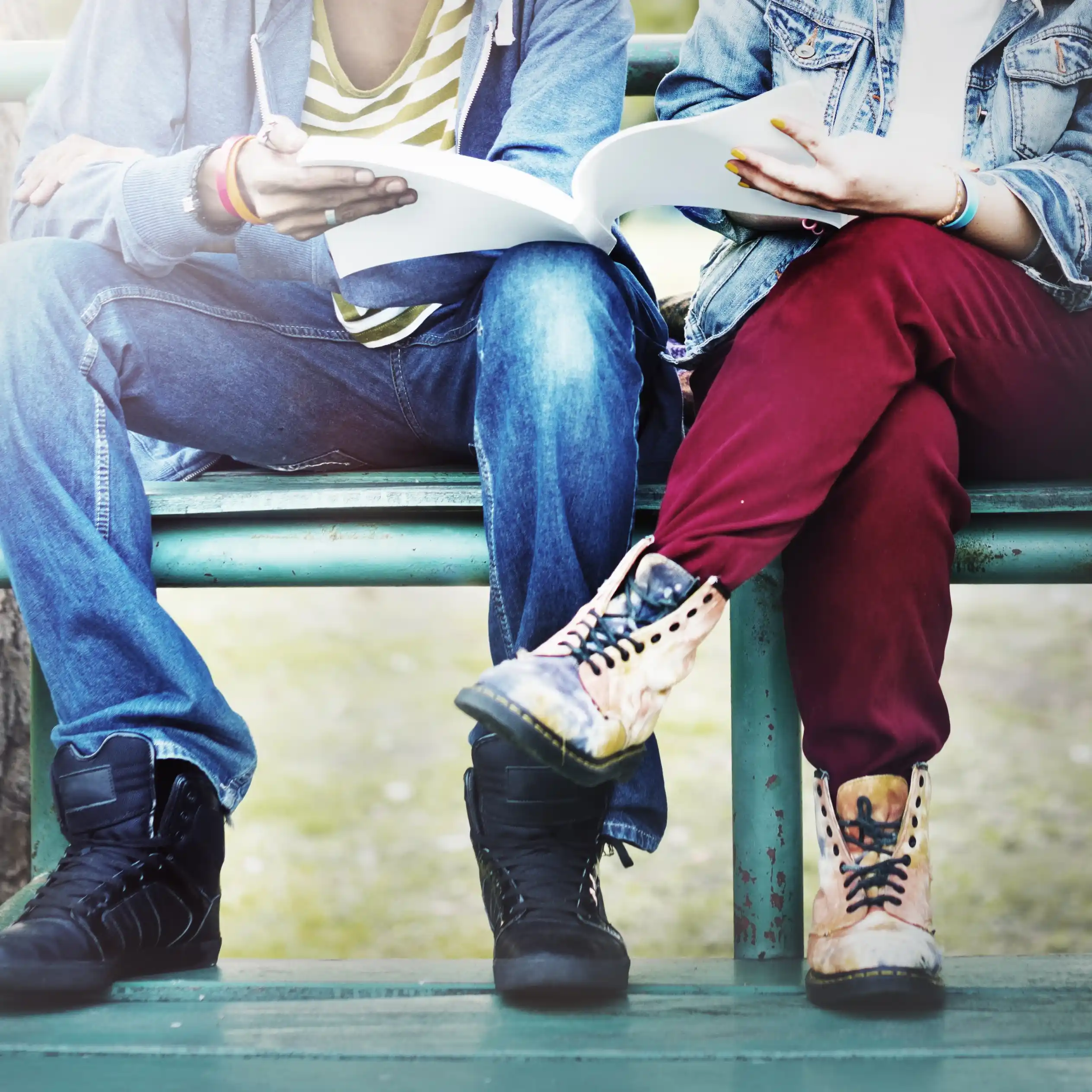 Dwie młode osoby siedzące na ławce na zewnątrz, czytające razem książkę lub notatki.