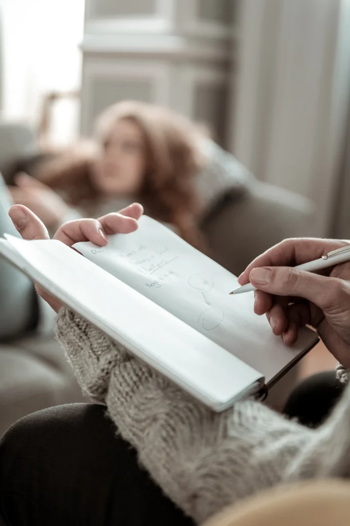 Zbliżenie na osobę robiącą notatki w zeszycie podczas sesji terapeutycznej, w tle rozmyta postać leżąca na kanapie